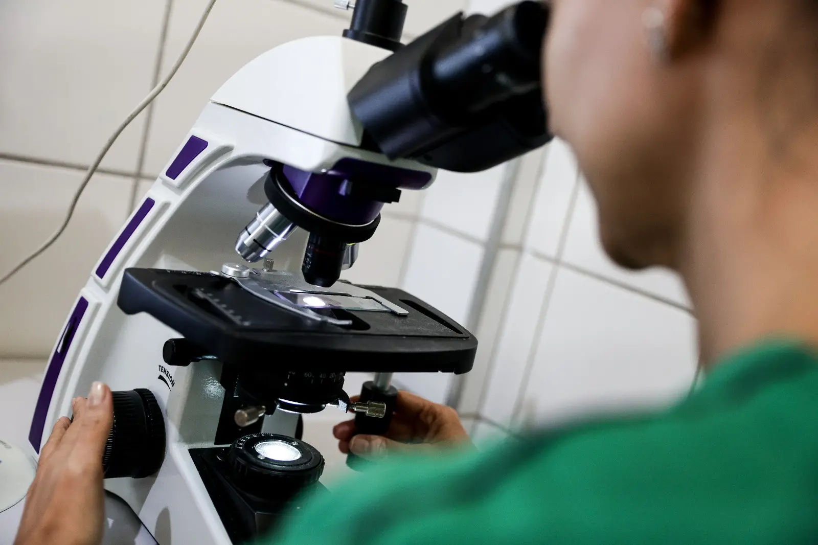 Uma profissional de saúde, vestindo uma camisa verde, está utilizando um microscópio óptico com um corpo principal branco e detalhes em roxo. Suas mãos estão ajustando o foco e a lâmina na platina do equipamento, realizando uma análise laboratorial.