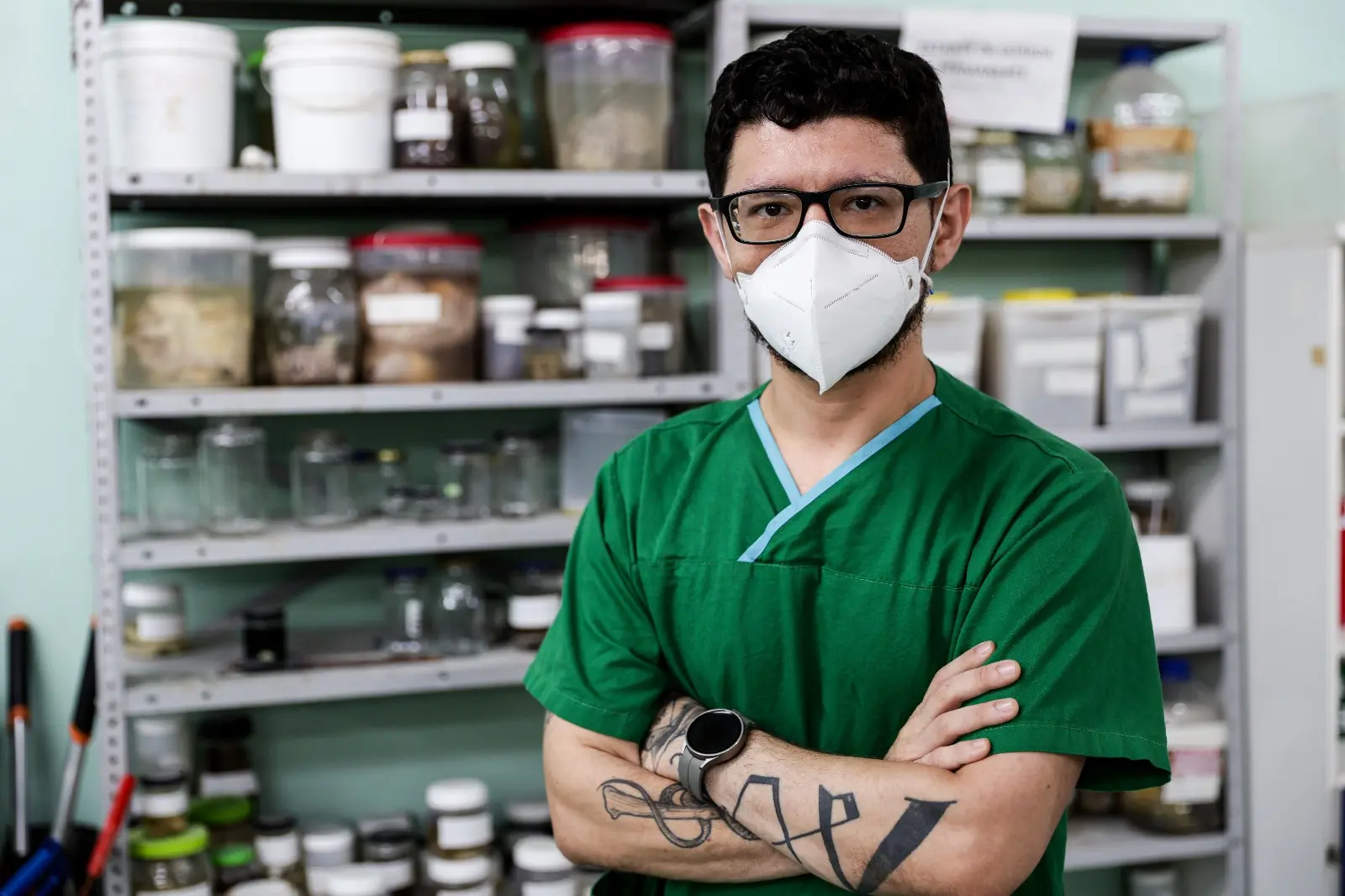 Um médico, identificado como Sami Gadelha, está em pé em um laboratório, vestindo uma blusa verde e uma máscara de proteção branca. Ele tem os braços cruzados, e no fundo há estantes repletas de potes e amostras.