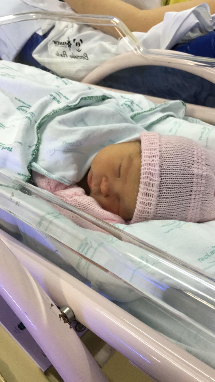 Foto vertical de um bebê recém-nascido dormindo. O bebê está deitado de lado em um berço hospitalar com laterais transparentes, usando uma touca e uma manta de tricô rosa. O bebê está coberto com lençóis brancos, e a pele amarelada sugere icterícia neonatal.