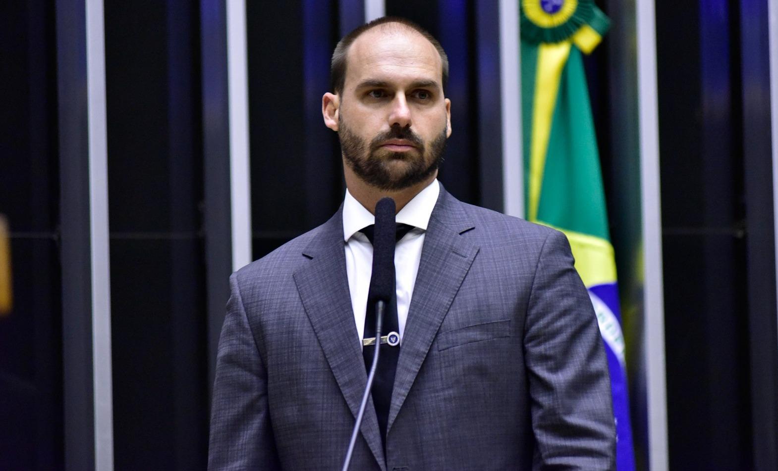 Eduardo Bolsonaro é um homem de meia idade, calvo e de barba rala. Na foto, ele usa terno cinza, camisa social branca e gravata preta.