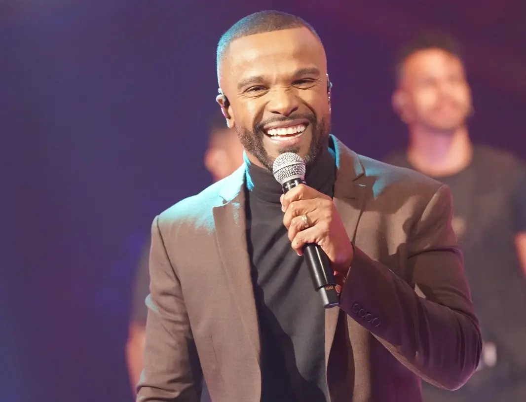Alexandre Pires é um homem negro de meia idade, careca e de barba rala. Na foto, ele usa terno marrom e camisa de gola alta da mesma cor. Está cantando.