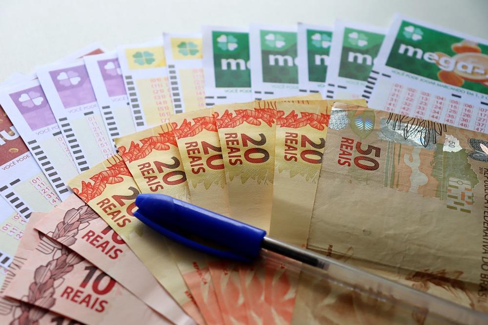 Loterias da Caixa Econômica Federal e cédulas de 20, de 10 e de 50 reais dispostas sobre uma mesa com uma caneta azul ao lado.