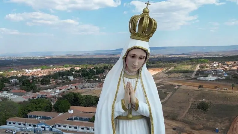Imagem de um grande monumento de Nossa Senhora, com manto branco e detalhes dourados, coroada e com as mãos postas em oração. A estátua aparece em primeiro plano, enquanto ao fundo há uma vista aérea de uma cidade, com casas, ruas, áreas verdes e um céu azul com nuvens dispersas.