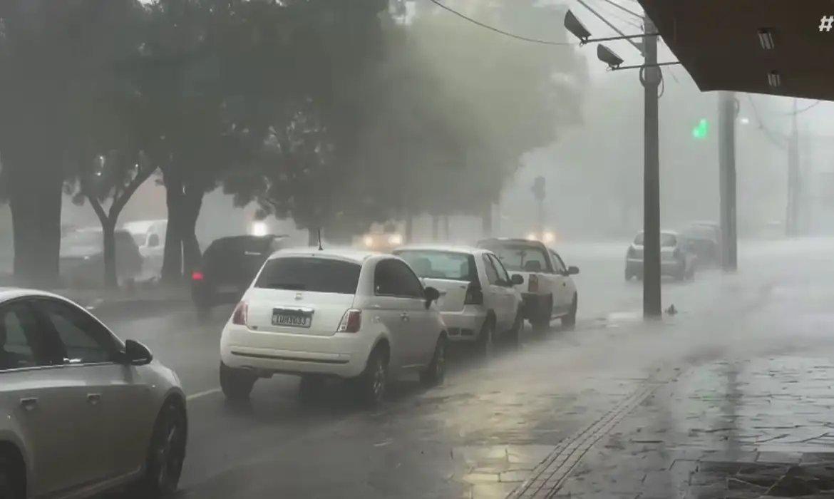 Rua urbana sob forte chuva e vento, com baixa visibilidade. Vários carros estão parados ou seguindo lentamente, enquanto a água se acumula no asfalto. Árvores ao fundo balançam com a intensidade da tempestade e a iluminação pública aparece difusa devido à neblina formada pela chuva. Imagem usada em matéria sobre alerta vermelho do Inmet.
