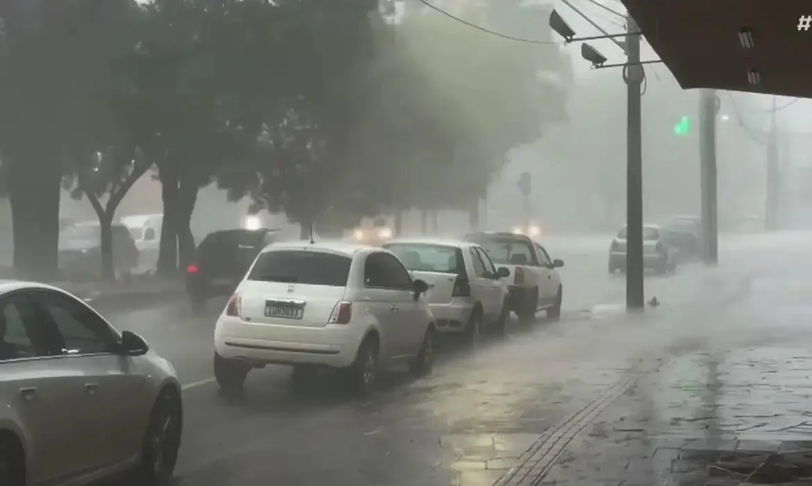 Rua urbana sob forte chuva e vento, com baixa visibilidade. Vários carros estão parados ou seguindo lentamente, enquanto a água se acumula no asfalto. Árvores ao fundo balançam com a intensidade da tempestade e a iluminação pública aparece difusa devido à neblina formada pela chuva. Imagem usada em matéria sobre alerta vermelho do Inmet.