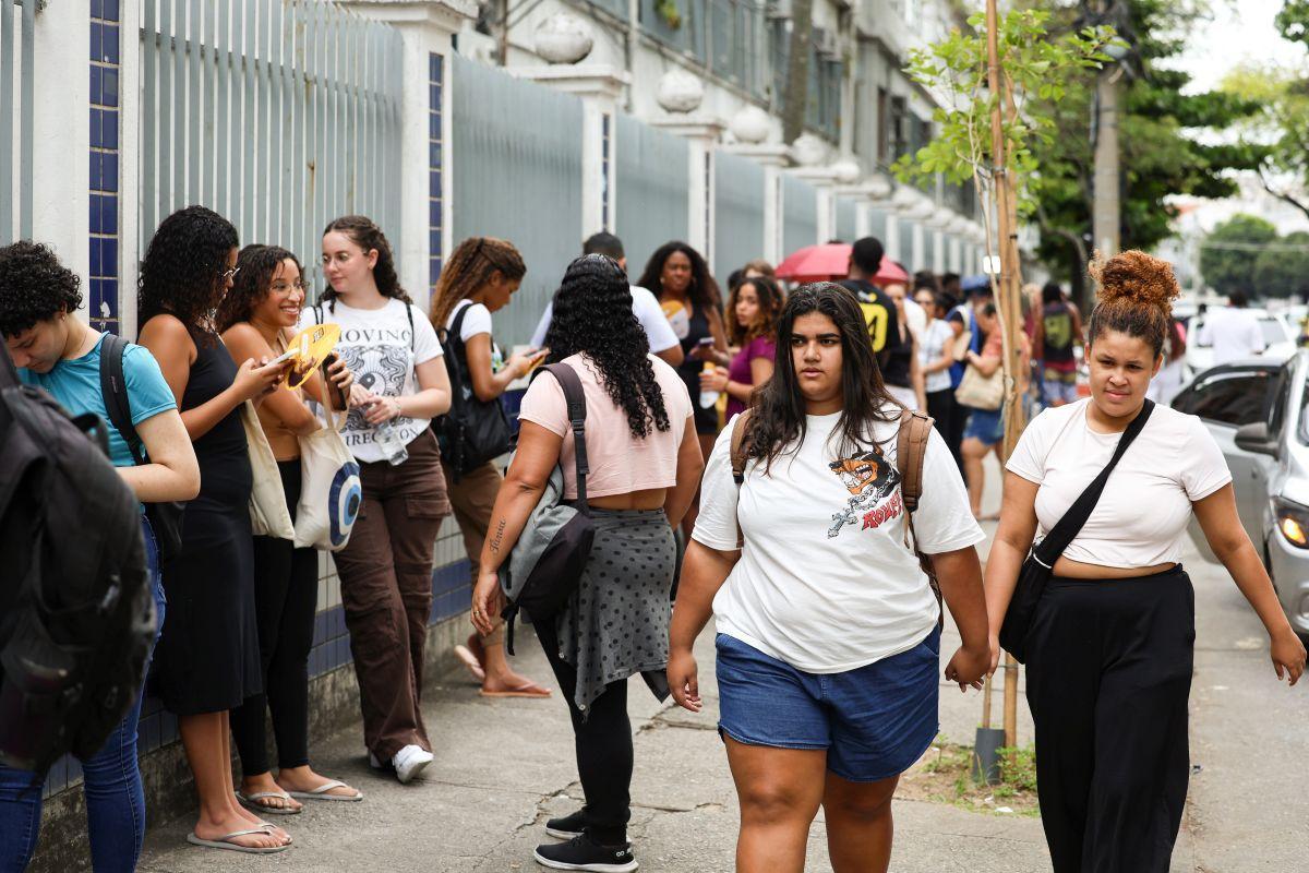 Grupo de pessoas reunidas em uma calçada ao lado de um muro com grades verticais e detalhes em azulejos azuis. Algumas pessoas estão em pé, usando mochilas e segurando celulares, enquanto outras caminham pela calçada. Há uma árvore jovem plantada próxima ao meio-fio e carros estacionados na rua ao fundo. A cena ocorre em frente a um prédio com fachada clara e janelas alinhadas. Imagem usada em matéria sobre Prova de Ciências da Natureza do Enem 2025.