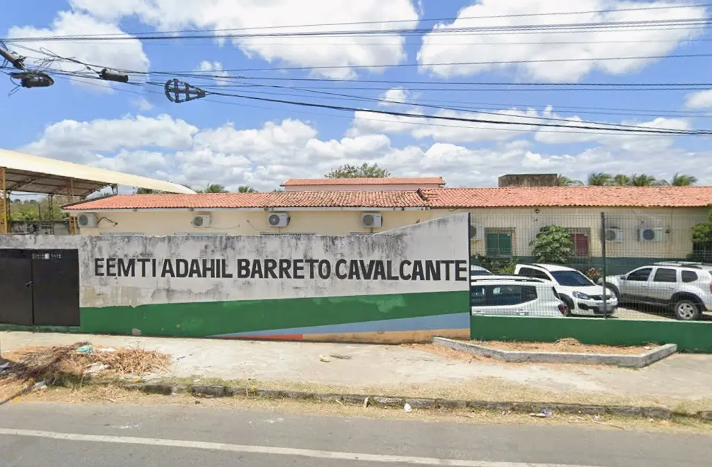 Fachada da EEMTI Adahil Barreto Cavalcante em Maracanaú, Ceará, com o nome da escola pintado em letras brancas sobre um muro listrado (verde, branco e azul). Parte de um complexo escolar de um andar é visível atrás do muro, e carros estão estacionados na frente.