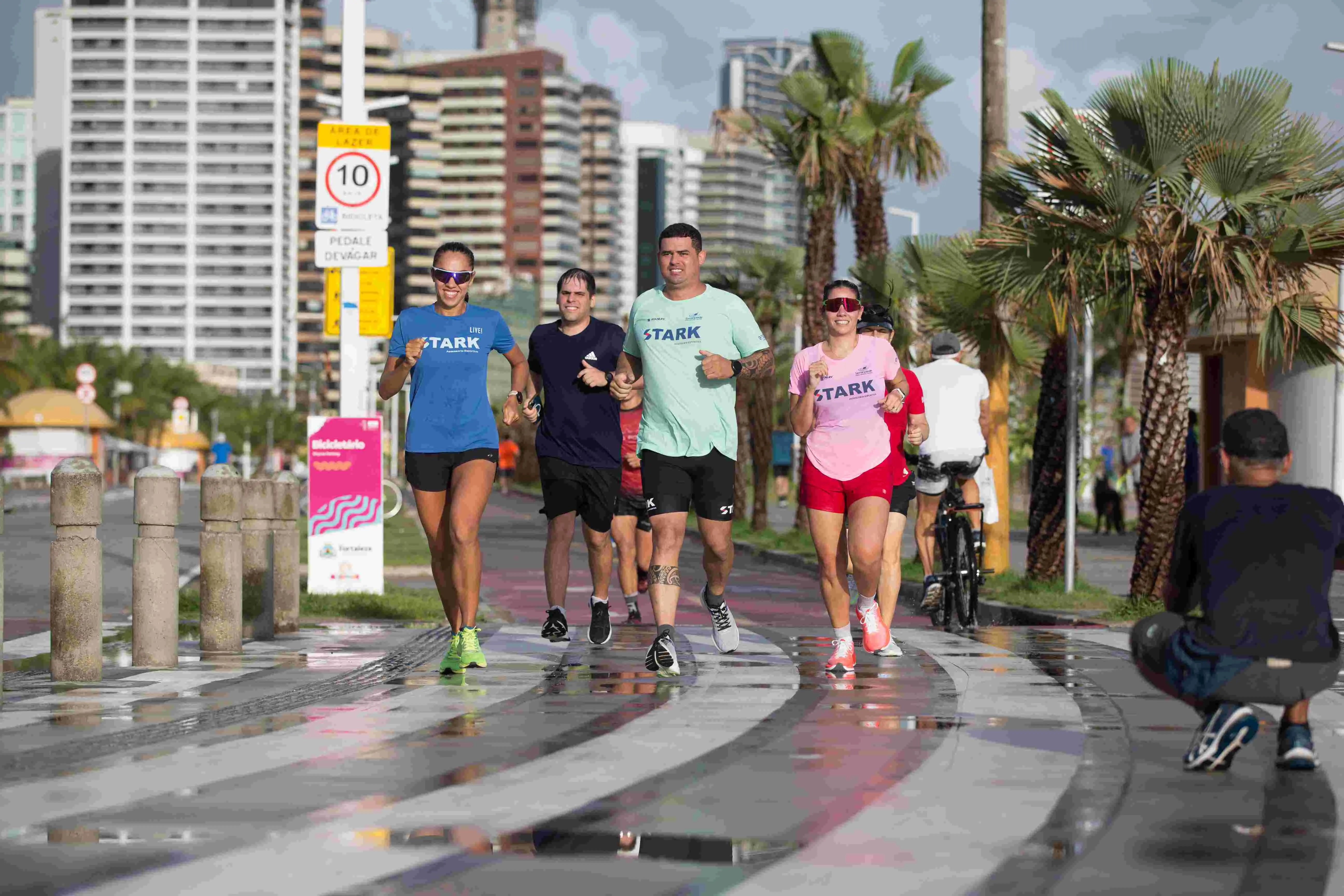 Quatro corredores (duas mulheres e dois homens), vestindo roupas esportivas de cores azul, preta, verde-água e rosa/vermelha, correm sorrindo pela calçada molhada da Avenida Beira Mar, em Fortaleza. Eles estão na faixa de pedestres/ciclistas, ladeados por coqueiros e prédios altos ao fundo, sob um céu parcialmente nublado. Um homem agachado no canto inferior direito parece estar tirando uma foto ou filmando a corrida.