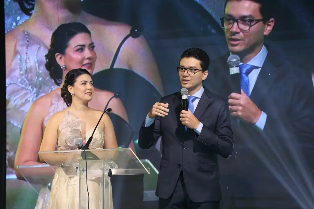 Apresentadores em palco durante evento formal, com bancada de acrílico, microfones e telão ao fundo exibindo suas imagens ampliadas. Um homem de terno e uma mulher em vestido claro conduzem a cerimônia