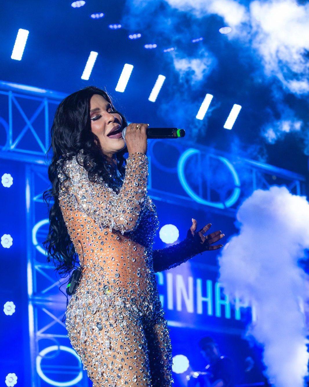 Artista feminina com microfone em show ao vivo, usando roupa cintilante e cenário iluminado.