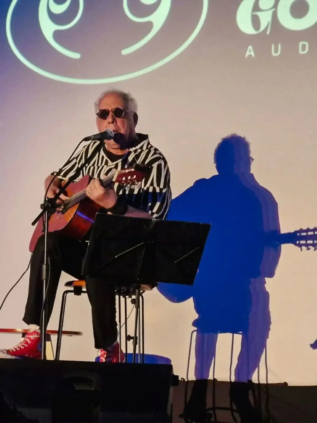 Músico sentado com violão, microfone e sapatos vermelhos, apresentando-se ao vivo sob um foco de luz que projeta sua sombra azul na parede.