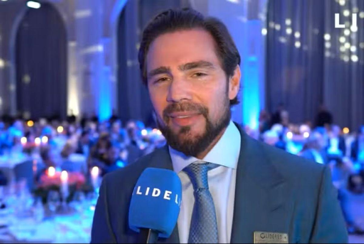 Foto de Daniel Vorcaro, de terno e gravata azul, falando em um microfone azul.