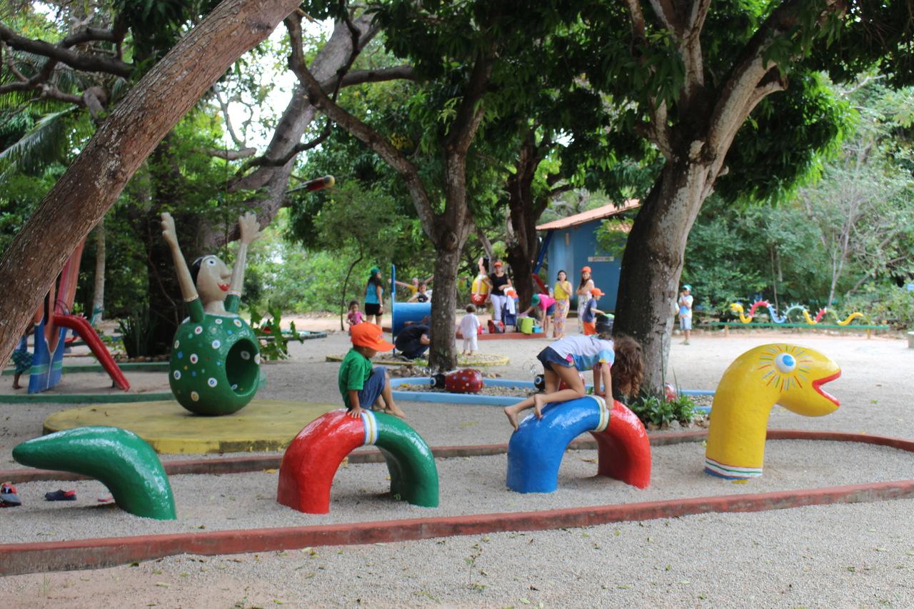 Crianças brincam em um parquinho externo com estruturas coloridas que são obras do mestre da cultura Dim Brinquedim sob a sombra de árvores grandes, com uma grande escultura de personagem verde ao fundo.