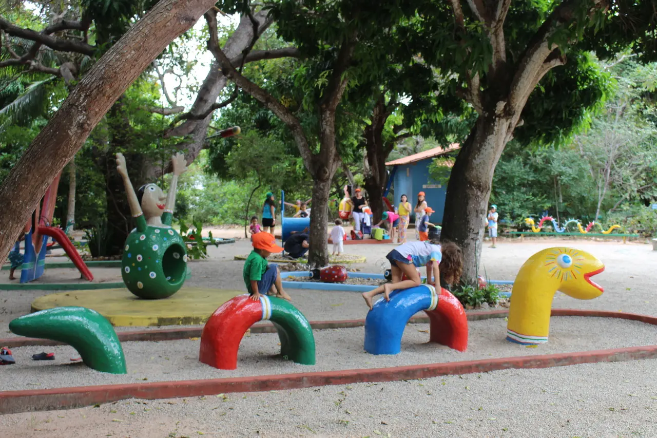 Crianças brincam em um parquinho externo com estruturas coloridas que são obras do mestre da cultura Dim Brinquedim sob a sombra de árvores grandes, com uma grande escultura de personagem verde ao fundo.