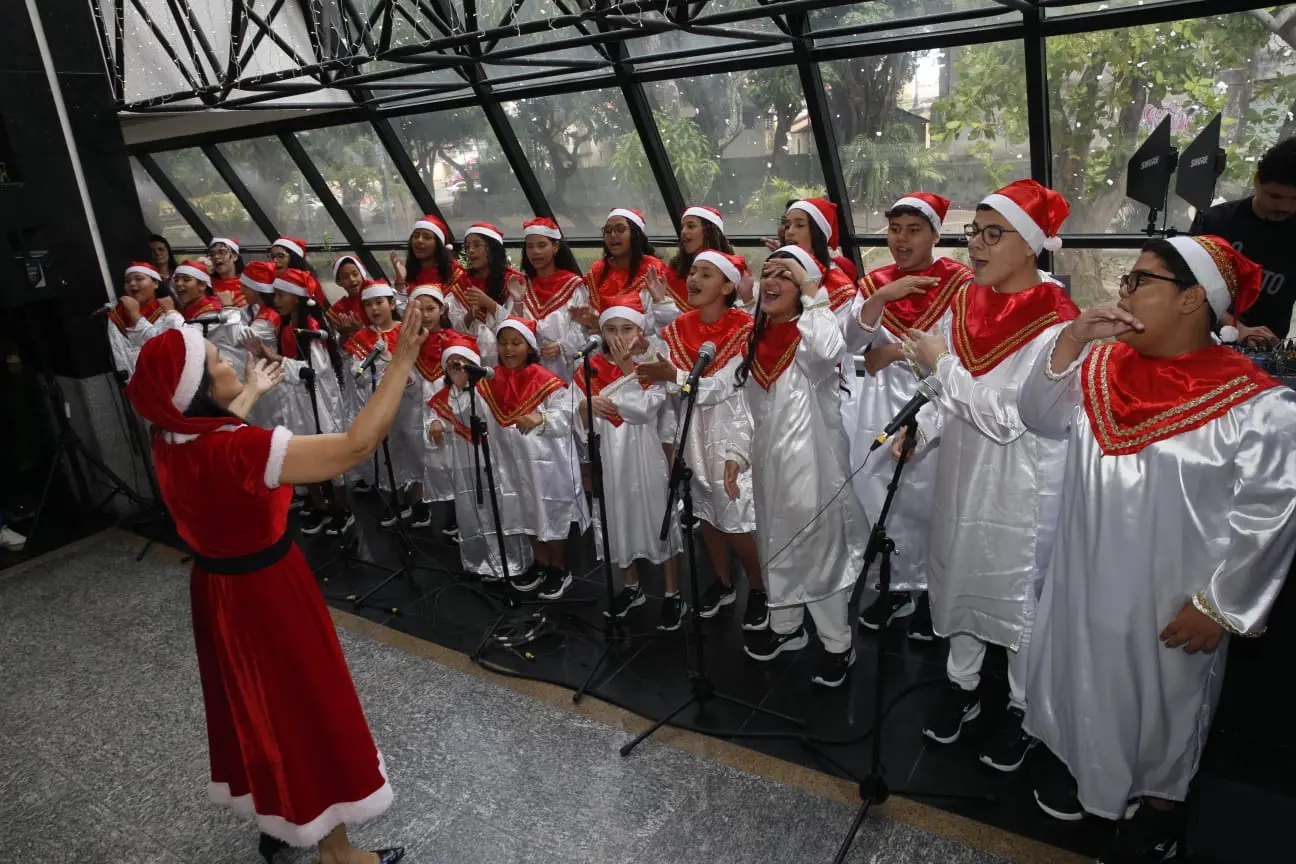 Crianças e jovens em um palco, com microfones à frente, performando em um coral de Natal, todos usando gorros de Papai Noel e uniformes brancos com detalhes em vermelho, sob a batuta de uma regente fantasiada.