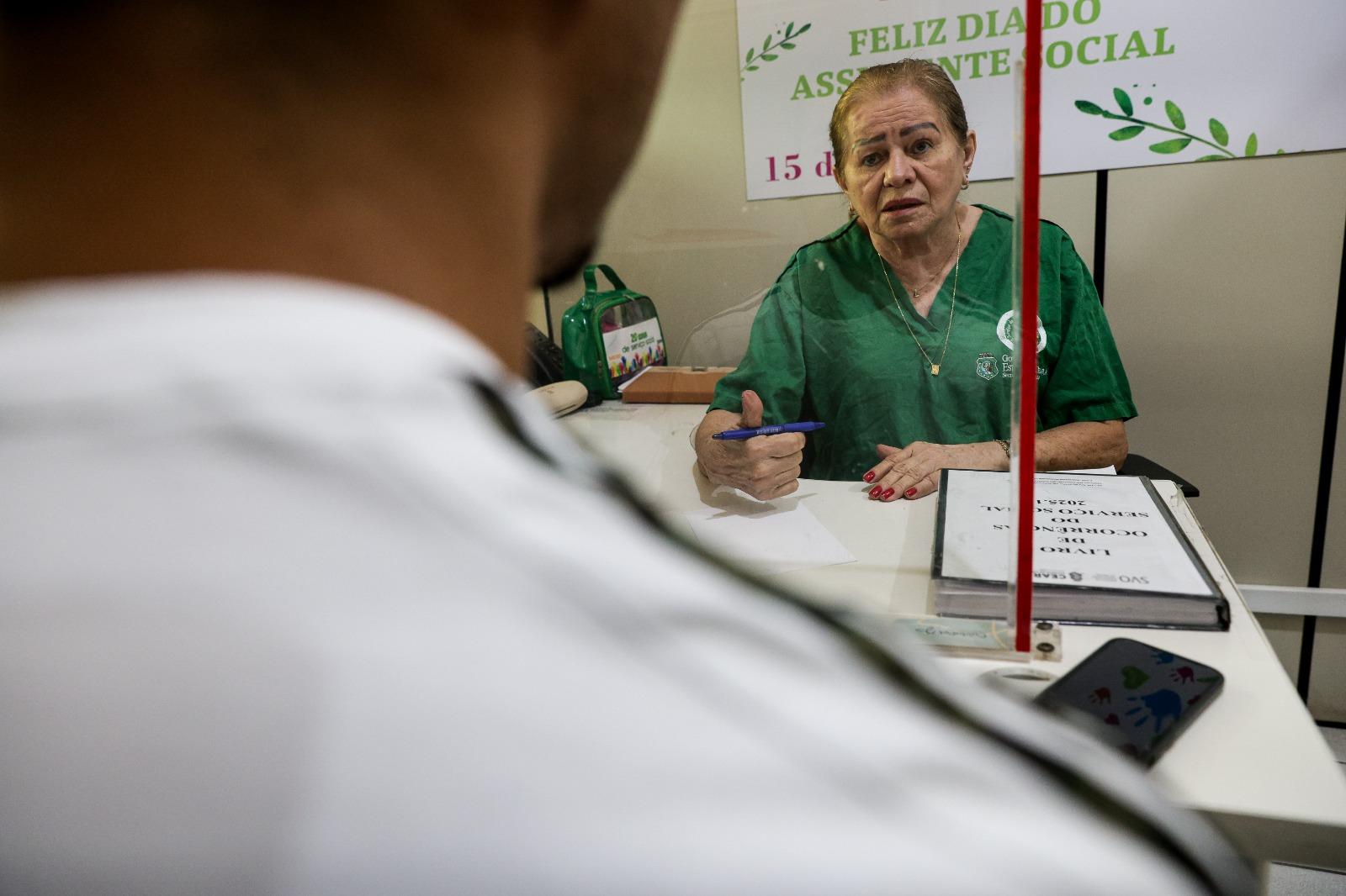 A assistente social Carmen Silva, uma mulher de meia-idade vestida com roupa verde, está sentada atrás de uma mesa, interagindo com uma pessoa que está fora do enquadramento. Ela gesticula com uma caneta e há uma placa de Feliz Dia do Assistente Social ao fundo.