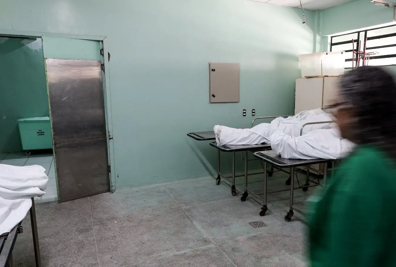 Uma sala de óbitos hospitalar com paredes verde-claras, onde se veem dois corpos cobertos por lençóis brancos em macas de transporte. Uma funcionária de scrubs verdes, parcialmente fora de foco, caminha no primeiro plano, à direita.