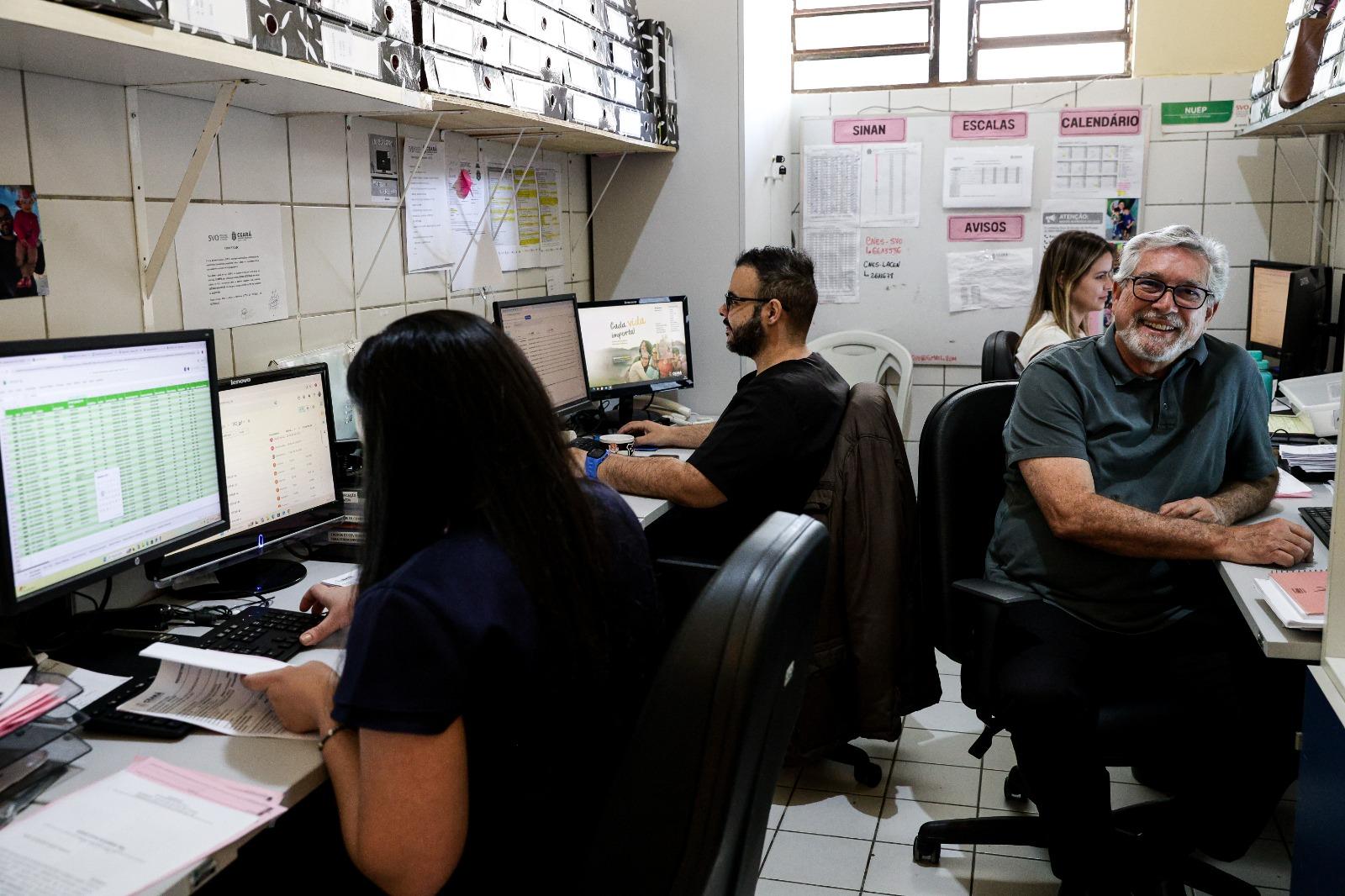 Pessoas trabalhando em um escritório, aparentemente administrativo de uma unidade de saúde (SVO). O médico José Rubens, um homem mais velho de camisa polo escura e óculos, sorri enquanto está sentado a uma mesa, olhando para a câmera. Outras pessoas estão focadas em seus monitores de computador.