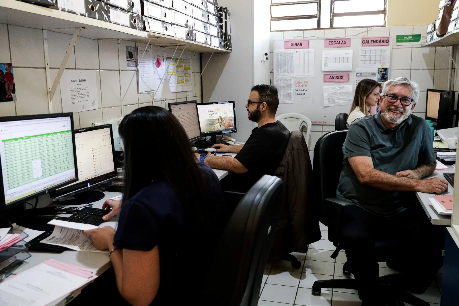 Pessoas trabalhando em um escritório, aparentemente administrativo de uma unidade de saúde (SVO). O médico José Rubens, um homem mais velho de camisa polo escura e óculos, sorri enquanto está sentado a uma mesa, olhando para a câmera. Outras pessoas estão focadas em seus monitores de computador.