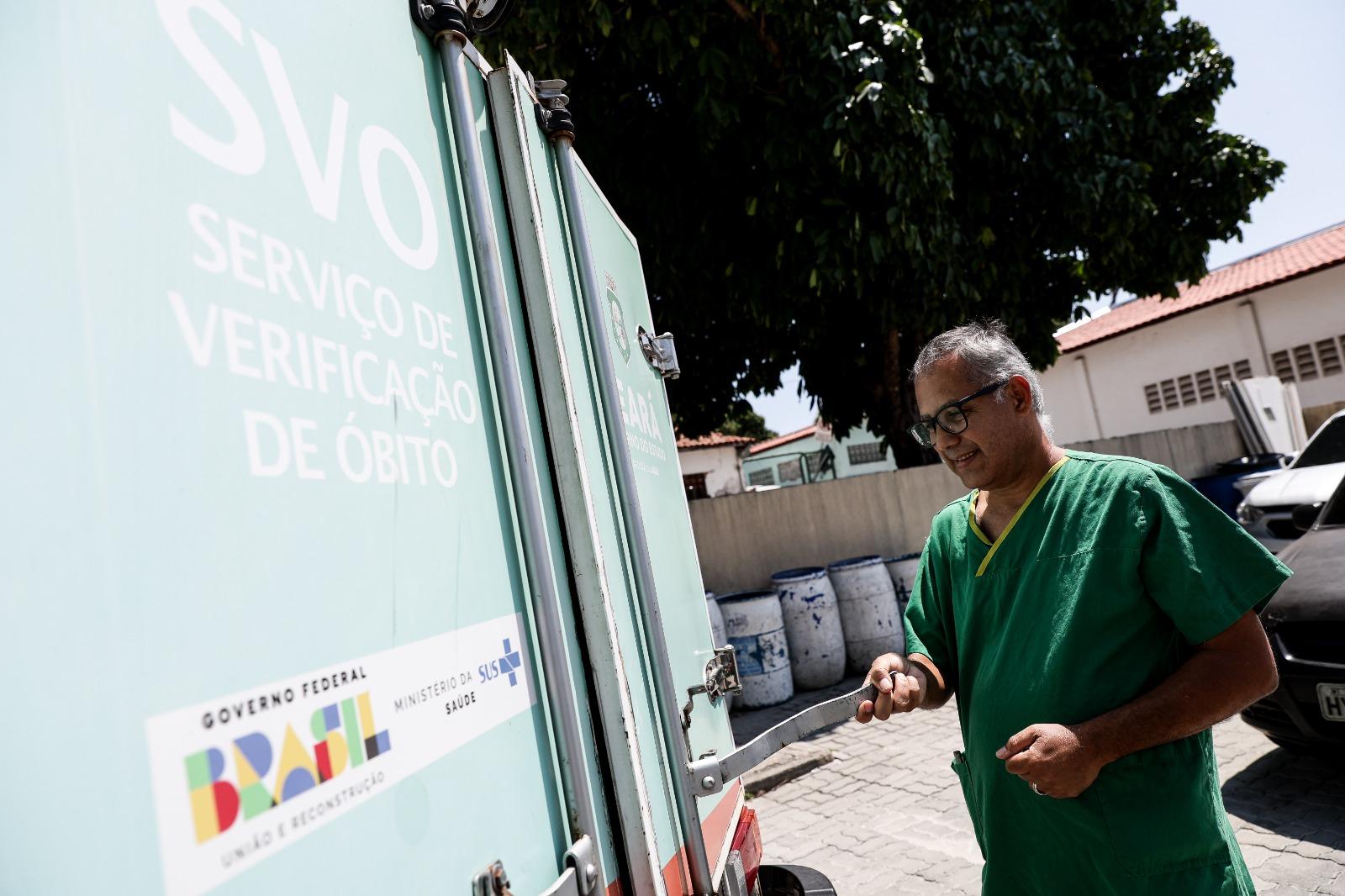 Um homem de scrubs verdes, identificado como Jair Cleudon, está ao ar livre, abrindo a porta traseira de um veículo do Serviço de Verificação de Óbito (SVO). A porta tem o logo do Governo Federal/Ministério da Saúde e a identificação do serviço.