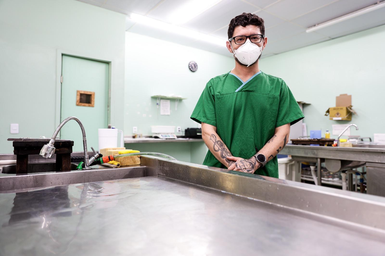 Um médico, identificado como Sami Gadelha, trajando scrubs verdes e máscara, está em um ambiente hospitalar que parece ser uma sala de autópsia ou necrotério, com mesas de aço inoxidável em primeiro plano. Ele tem tatuagens nos antebraços e usa óculos.