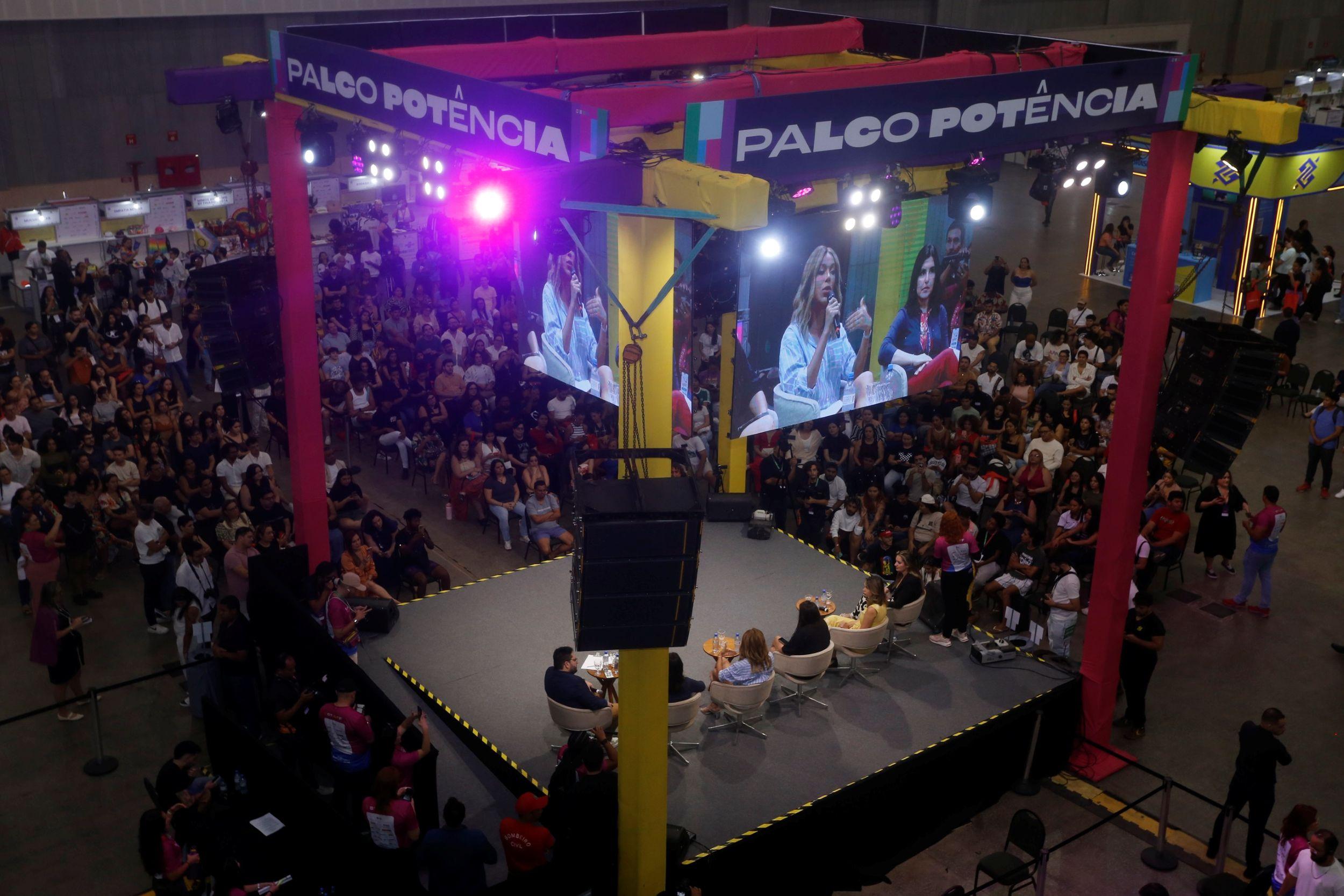 Vista superior de um grande evento com o 'Palco Potência' no centro, onde um painel de debate acontece diante de uma plateia numerosa e telões exibem imagens de mulheres.