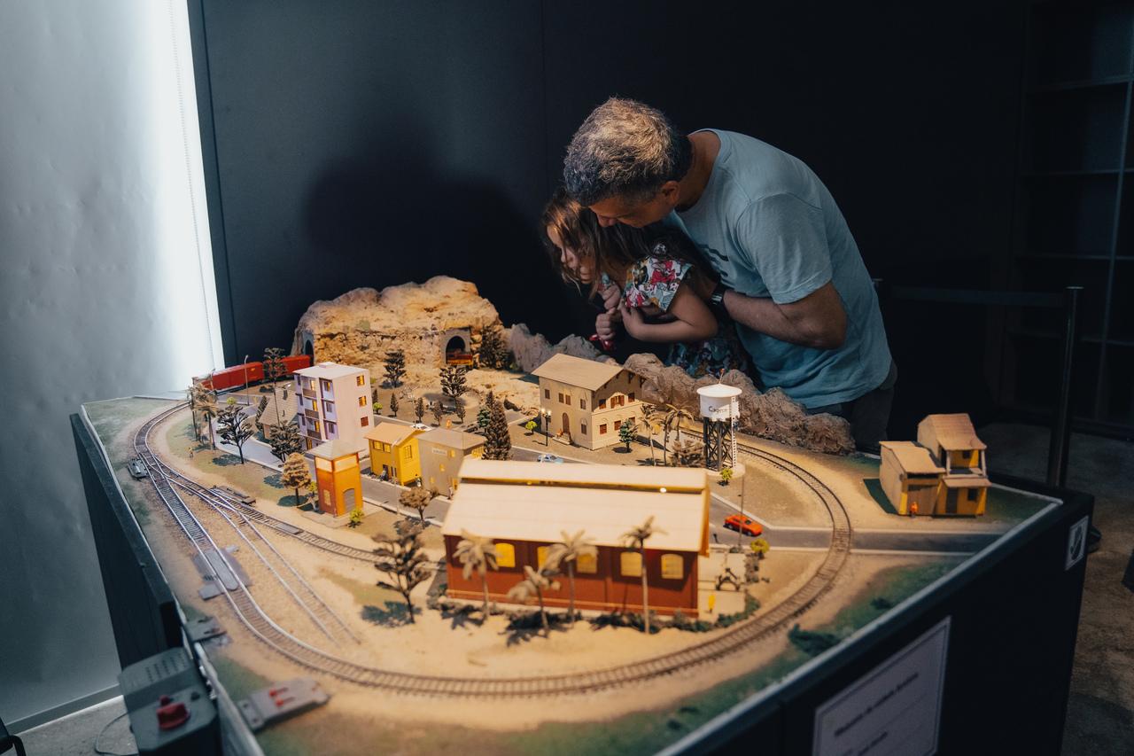 Um adulto e uma criança observam atentamente uma maquete de ferrovia detalhada com casas e paisagens, com foco nas miniaturas e trilhos.
