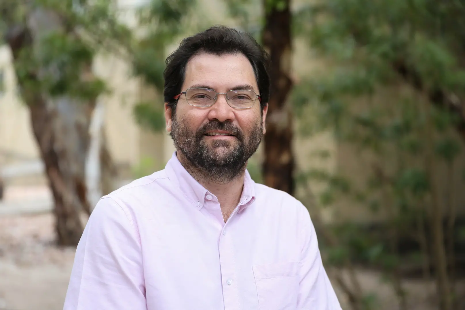 Um retrato de Antonio Silva Lima Neto, um homem barbudo com óculos, que usa uma camisa social rosa clara. Ele está sorrindo levemente e a foto foi tirada ao ar livre, com árvores e folhagem em segundo plano, ligeiramente desfocadas.