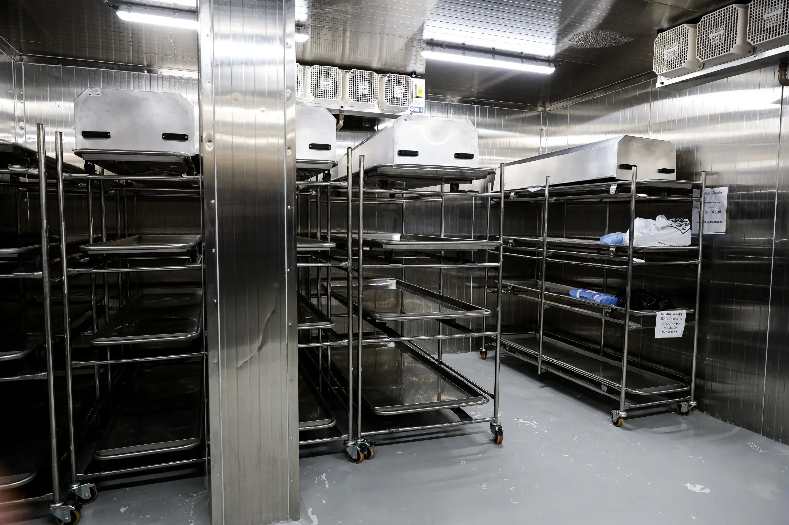 O interior de uma câmara fria de metal inoxidável, dentro do Serviço de Verificação de Óbito (SVO) de Fortaleza. A sala contém várias estantes metálicas de rodízios com bandejas, algumas vazias e outras com recipientes de plástico ou tampas metálicas que cobrem corpos.