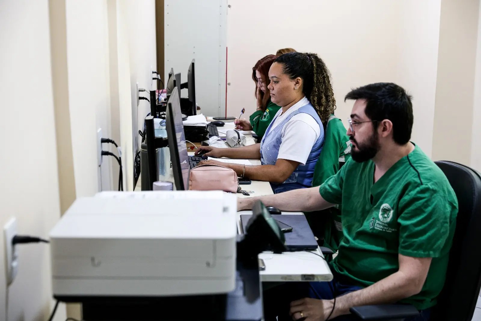 Uma cena de escritório onde três pessoas estão sentadas lado a lado, trabalhando em computadores. A pessoa em primeiro plano, à direita, é um homem barbudo vestindo scrubs verdes e óculos no rosto, focado no seu trabalho. As outras duas são mulheres que atuam na equipe de regulação do SVO Ceará.