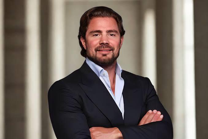 Homem barbudo de terno escuro e camisa azul sorri com os braços cruzados, em ambiente profissional com colunas.