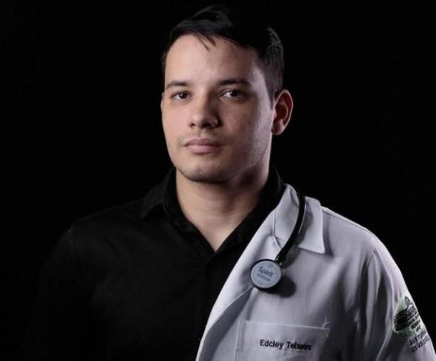 Edcley é um jovem de cabelo preto. Na foto, com fundo preto, ele está sério e usa jaleco de médico.