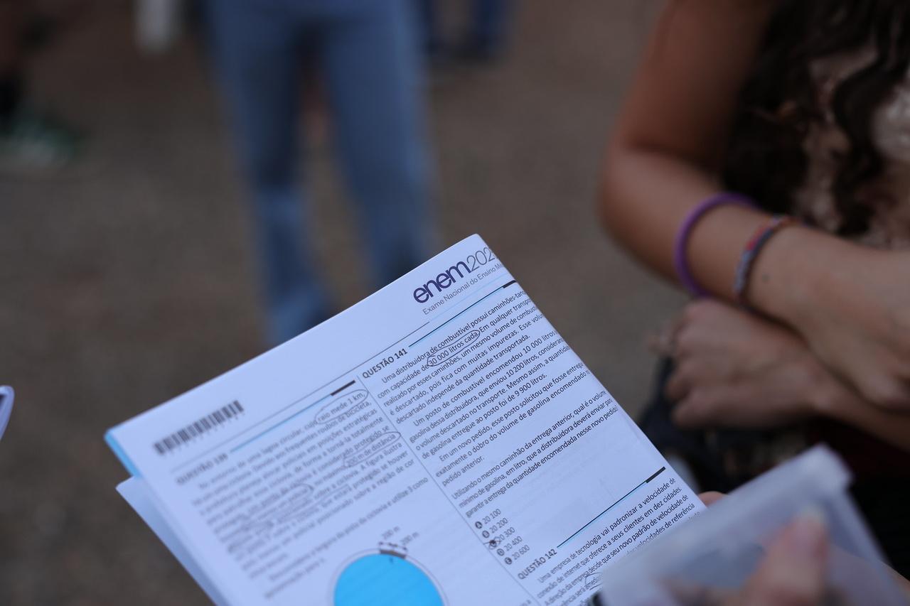Prova de vestibular com questões em branco. Ao fundo, em desfoque, pés de pessoas em pé.