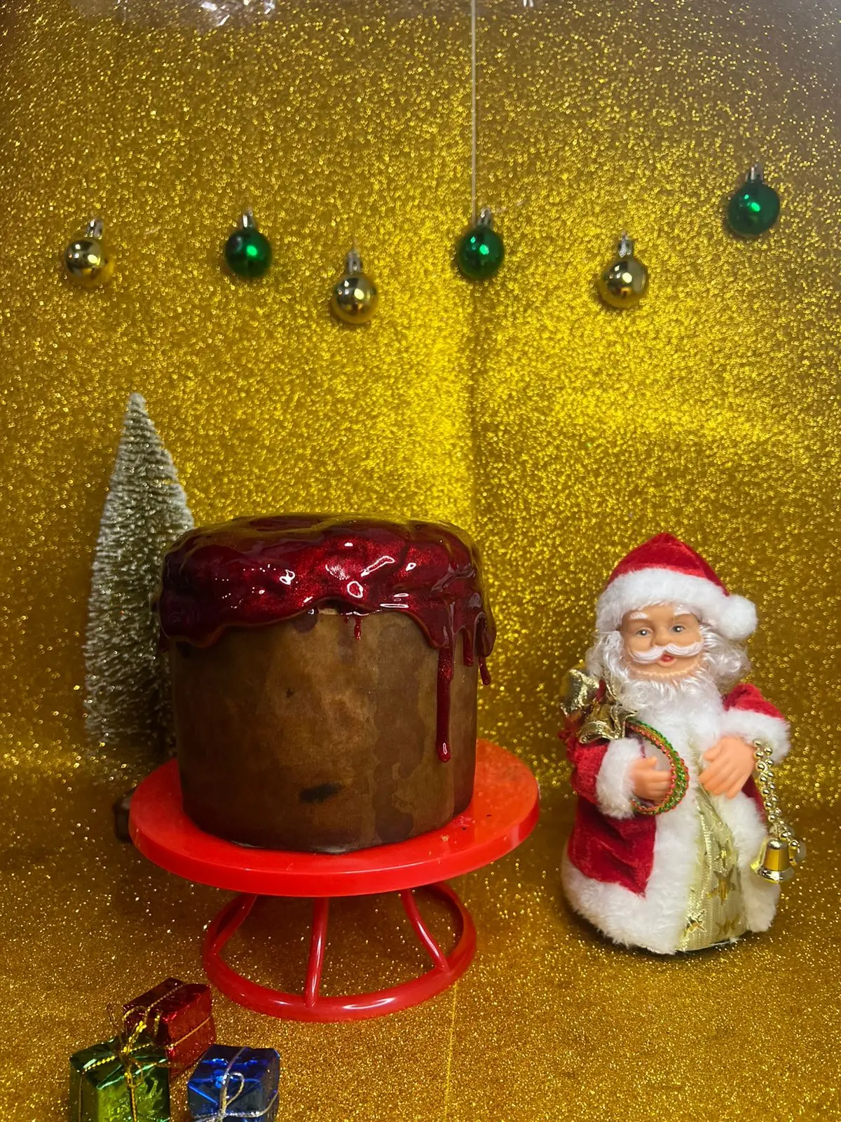 Panetone coberto com calda vermelha sobre um suporte vermelho, decorado com fundo dourado brilhante, pequenas bolas verdes suspensas, árvore em miniatura e presentes coloridos ao lado de uma figura natalina.