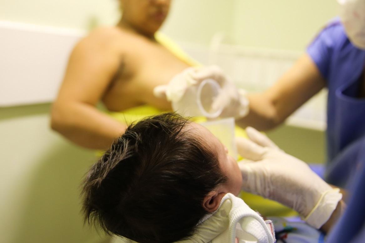 Uma cena focada no cuidado de um recém-nascido, provavelmente em um Banco de Leite Humano (como sugere o nome do arquivo, HGCC). Uma profissional de saúde, usando luvas e scrubs azuis, alimenta o bebê, que está deitado e tem a cabeça em primeiro plano. Em segundo plano, uma mãe com uma blusa amarela está sentada e sem camisa, sugerindo um momento de amamentação ou coleta de leite.