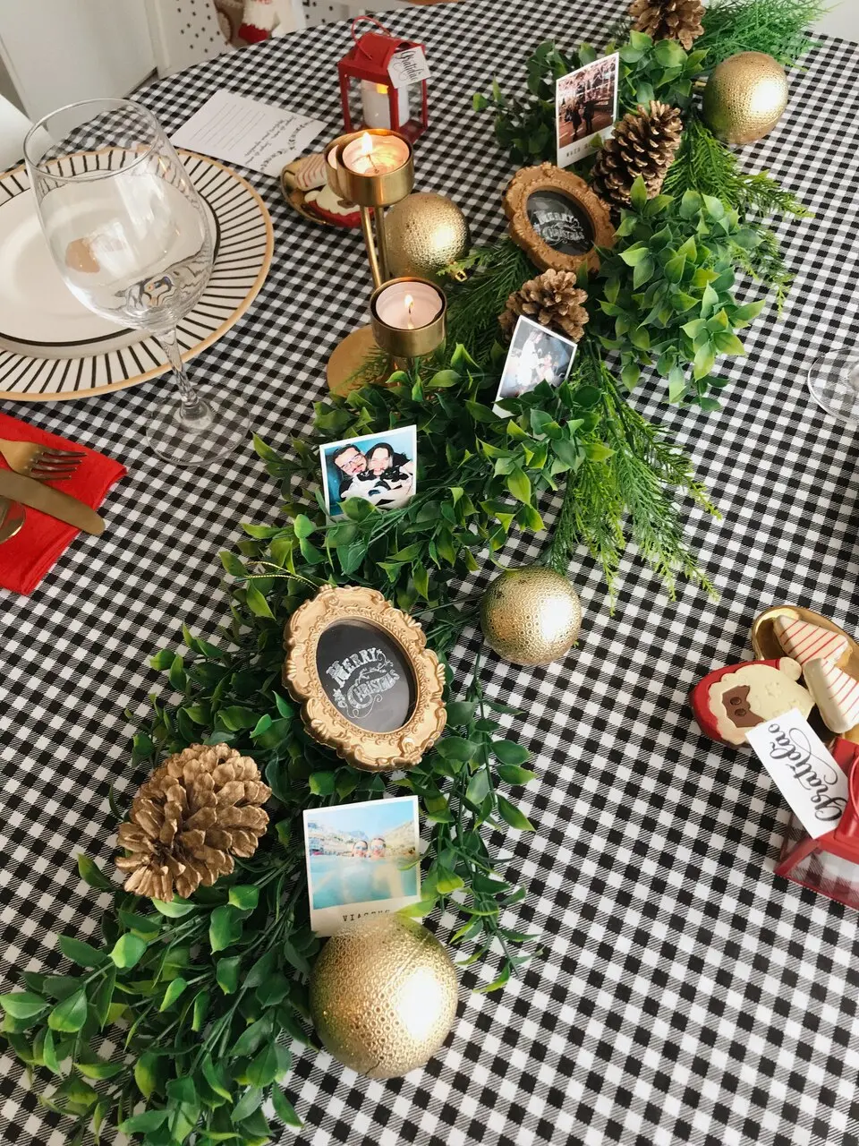 Uma mesa com toalha xadrez preta e branca está decorada para uma ocasião especial, com um centro de mesa que combina folhagens verdes, pinhas douradas e bolas decorativas douradas. Pequenas fotos pessoais e porta-retratos dourados em formato oval, alguns com a palavra 
