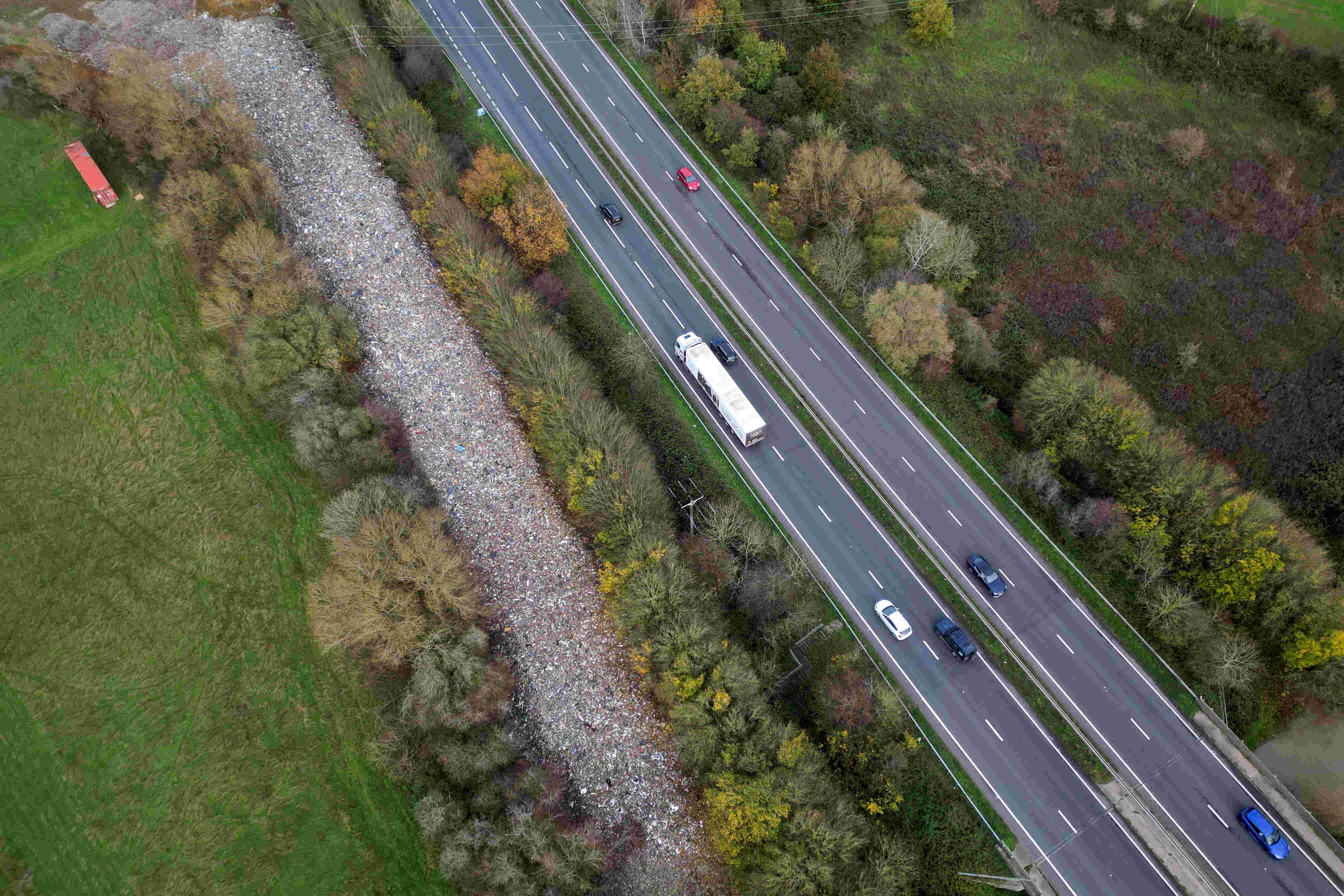 Imagem aérea mostra uma rodovia de pista dupla com tráfego moderado, incluindo carros e um caminhão branco, ladeada por áreas verdes e árvores. Ao lado da estrada, há uma extensa faixa coberta por entulho e resíduos, contrastando com o campo verde ao redor. As copas das árvores exibem tons variados de verde, marrom e amarelo, indicando estação outonal.