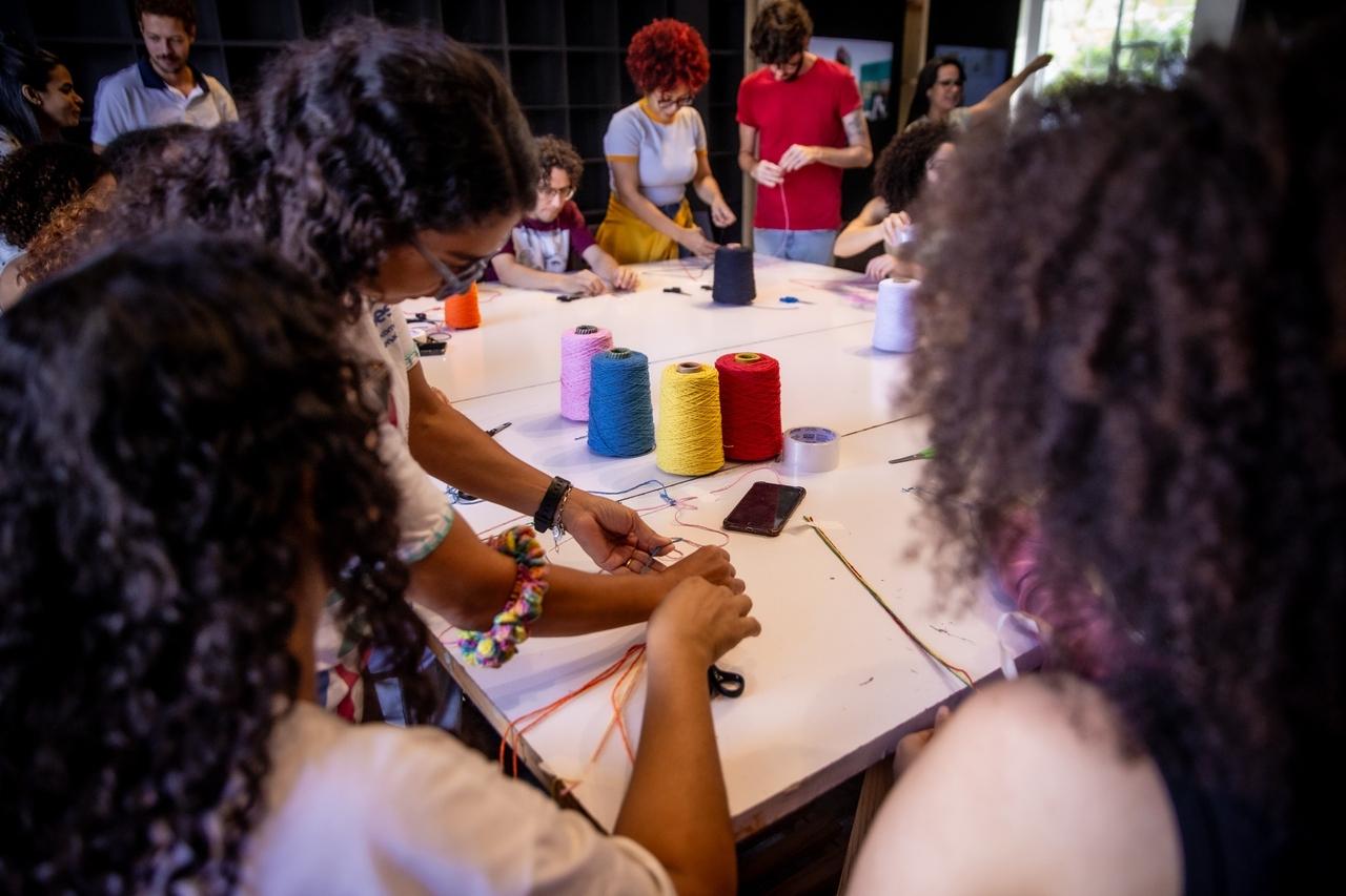 Pessoas em torno de uma mesa branca, fazendo artesanato com carretéis de linha coloridos e fios, como parte de uma atividade cultural em um equipamento de arte.