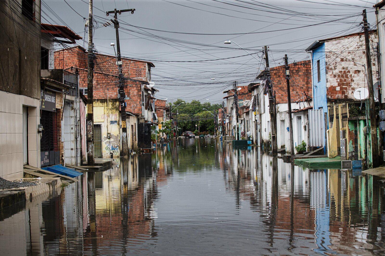 Vista de uma rua estreita com água da enchente refletindo as fachadas coloridas das casas em uma área urbana.