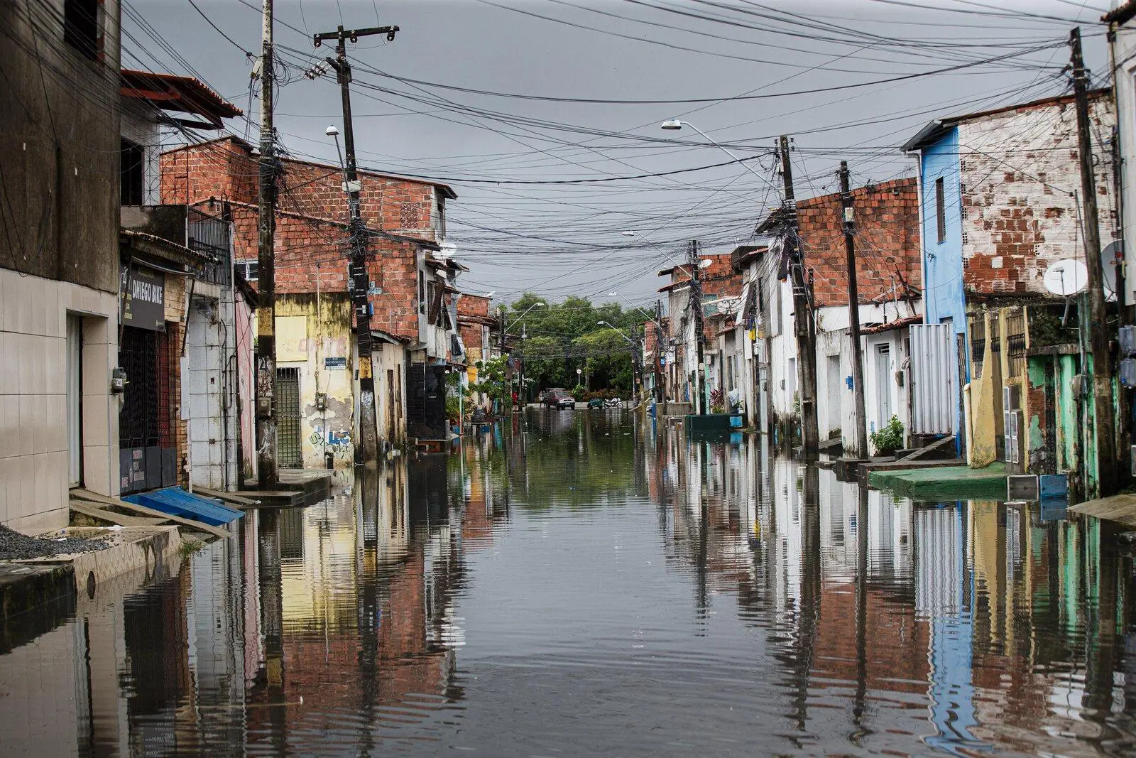 Vista de uma rua estreita com água da enchente refletindo as fachadas coloridas das casas em uma área urbana.