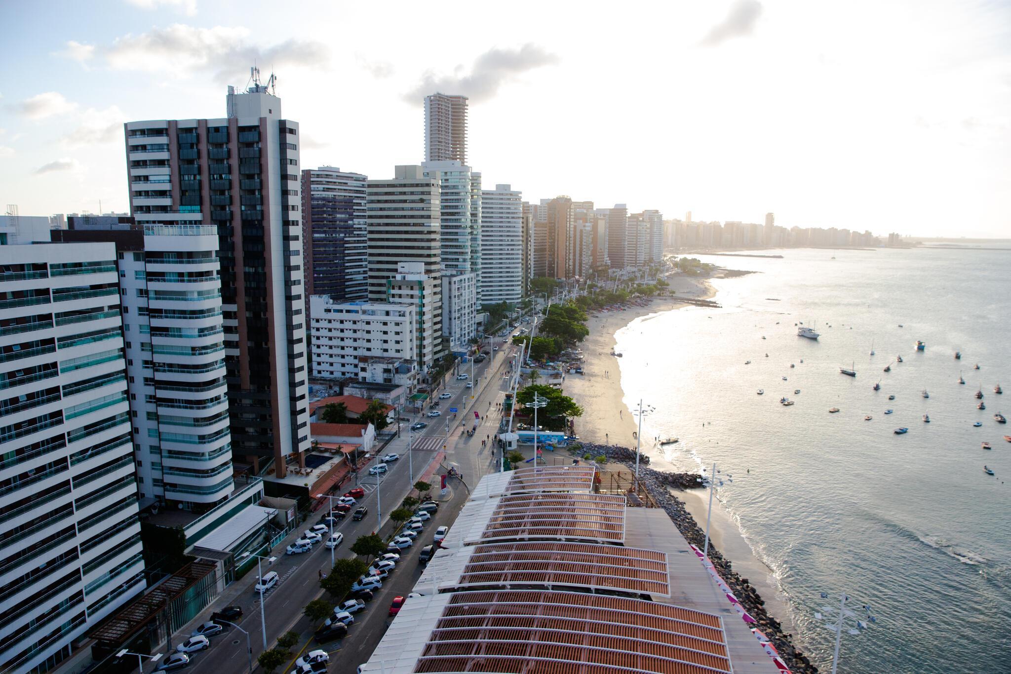 Imagem aérea da orla da Beira Mar, em Fortaleza. À esquerda, prédios. À direita, o mar.