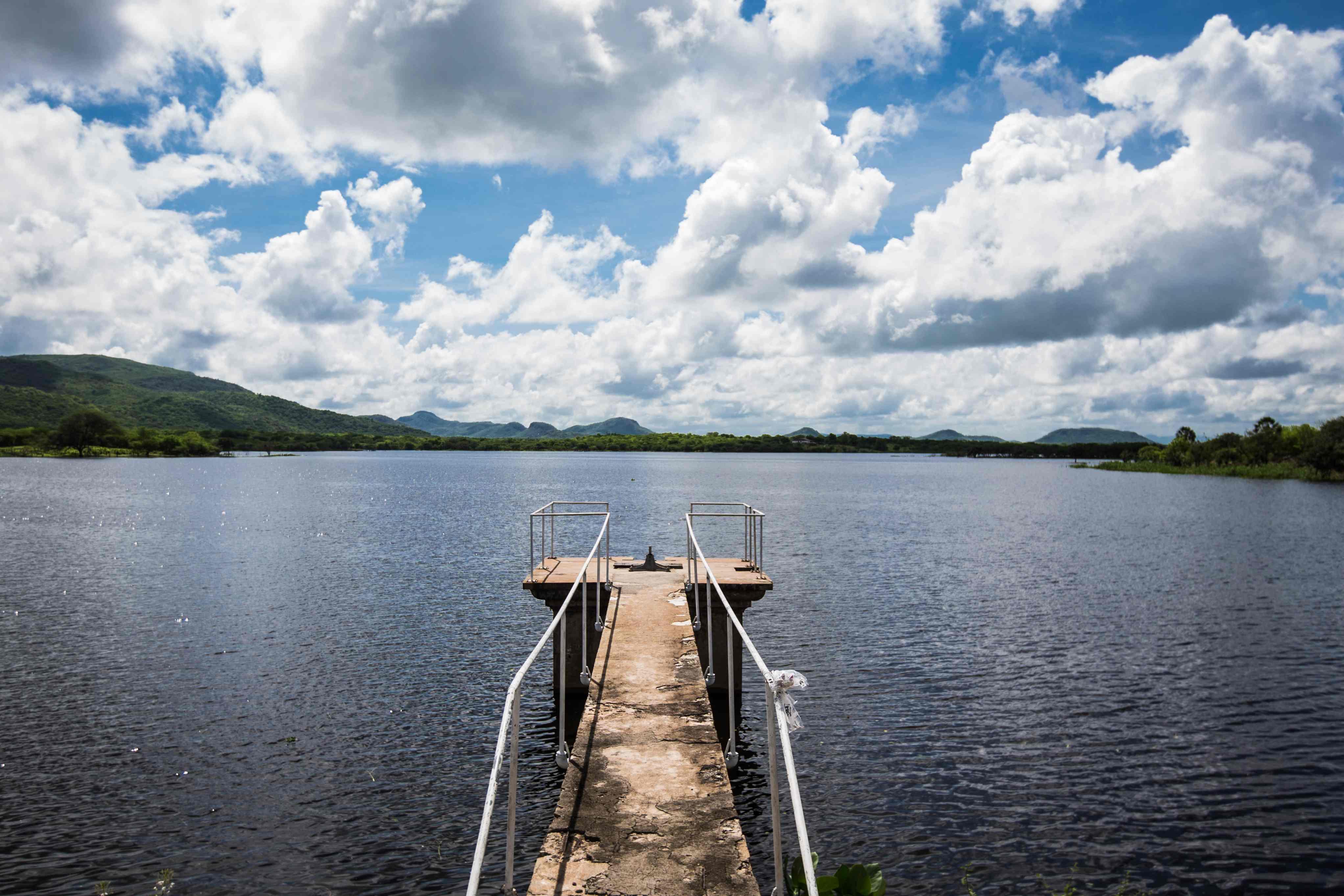 Vista do Açude de Patos, no Ceará. Um pequeno e estreito cais de concreto com corrimãos metálicos se estende sobre a água escura do açude. Ao fundo, o horizonte mostra morros cobertos por vegetação verde sob um céu azul com muitas nuvens brancas.