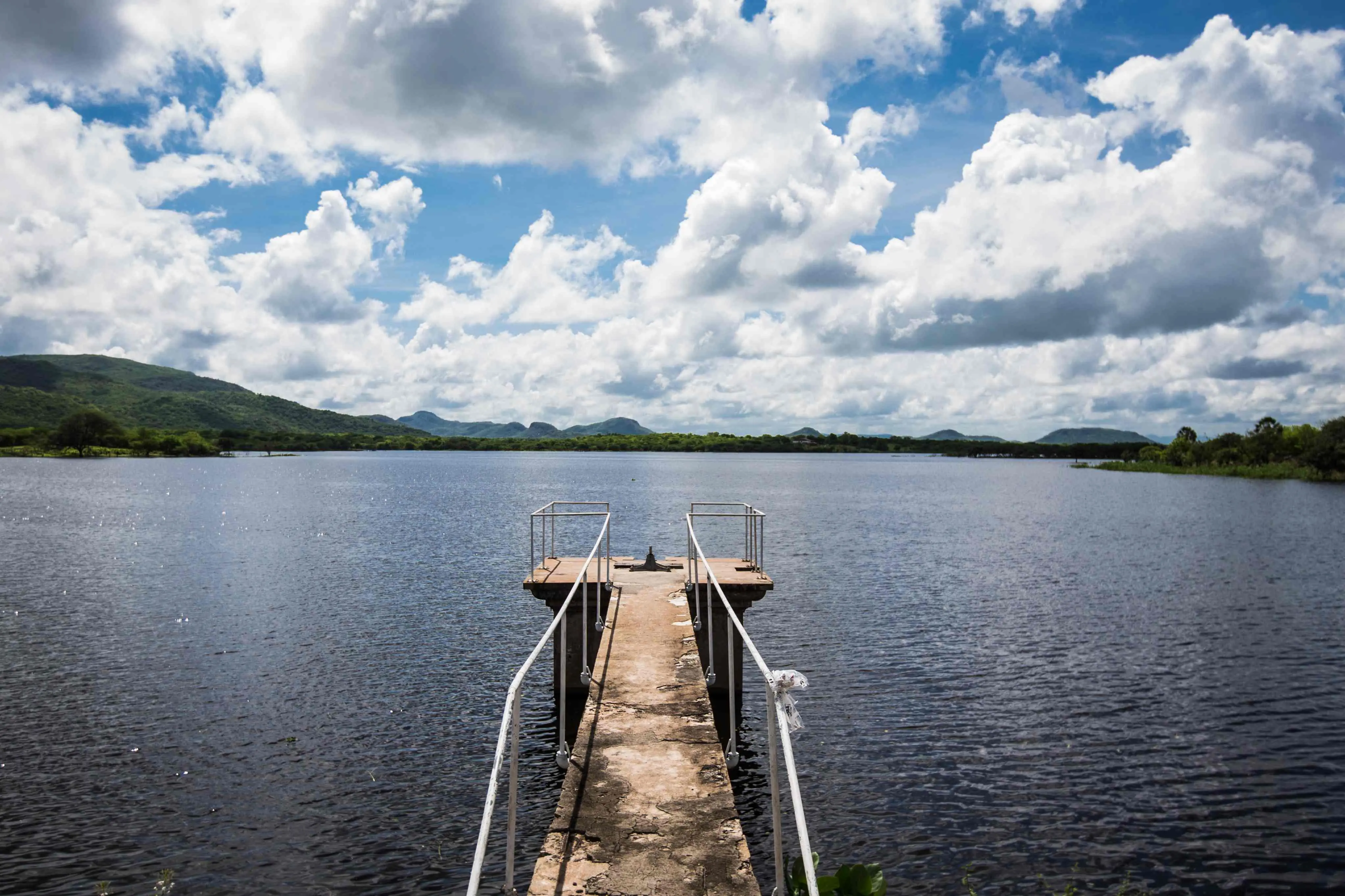 Vista do Açude de Patos, no Ceará. Um pequeno e estreito cais de concreto com corrimãos metálicos se estende sobre a água escura do açude. Ao fundo, o horizonte mostra morros cobertos por vegetação verde sob um céu azul com muitas nuvens brancas.