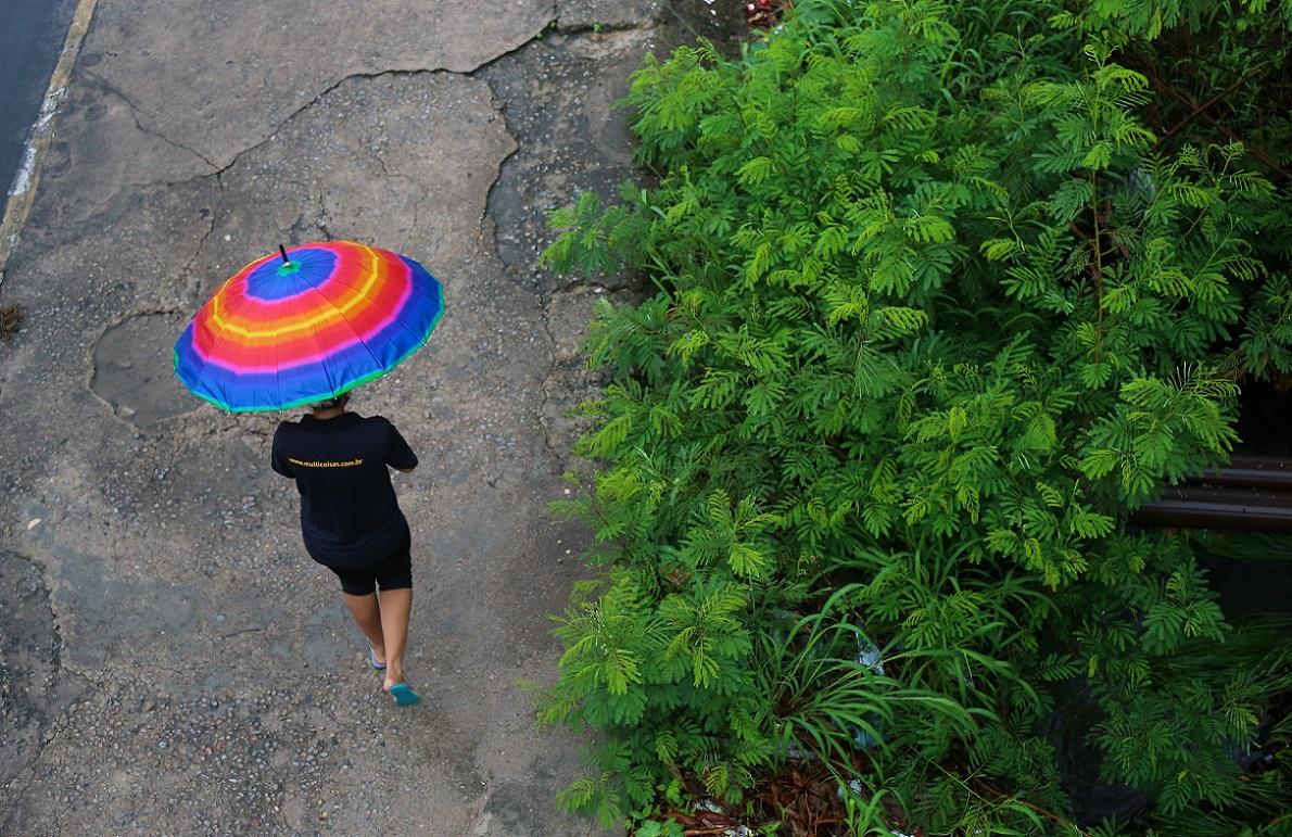 Uma vista aérea de uma mulher caminhando em uma área de concreto/asfalto rachado, provavelmente durante ou logo após a chuva. Ela usa uma camiseta preta e segura um guarda-chuva grande com as cores do arco-íris, que se destaca. À direita, há uma densa vegetação verde.