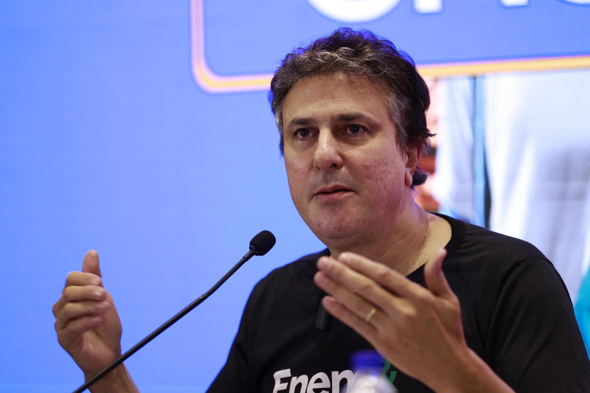 Homem com cabelo escuro e camiseta preta gesticula enquanto fala em um microfone em um evento. O homem é Camilo Santana.