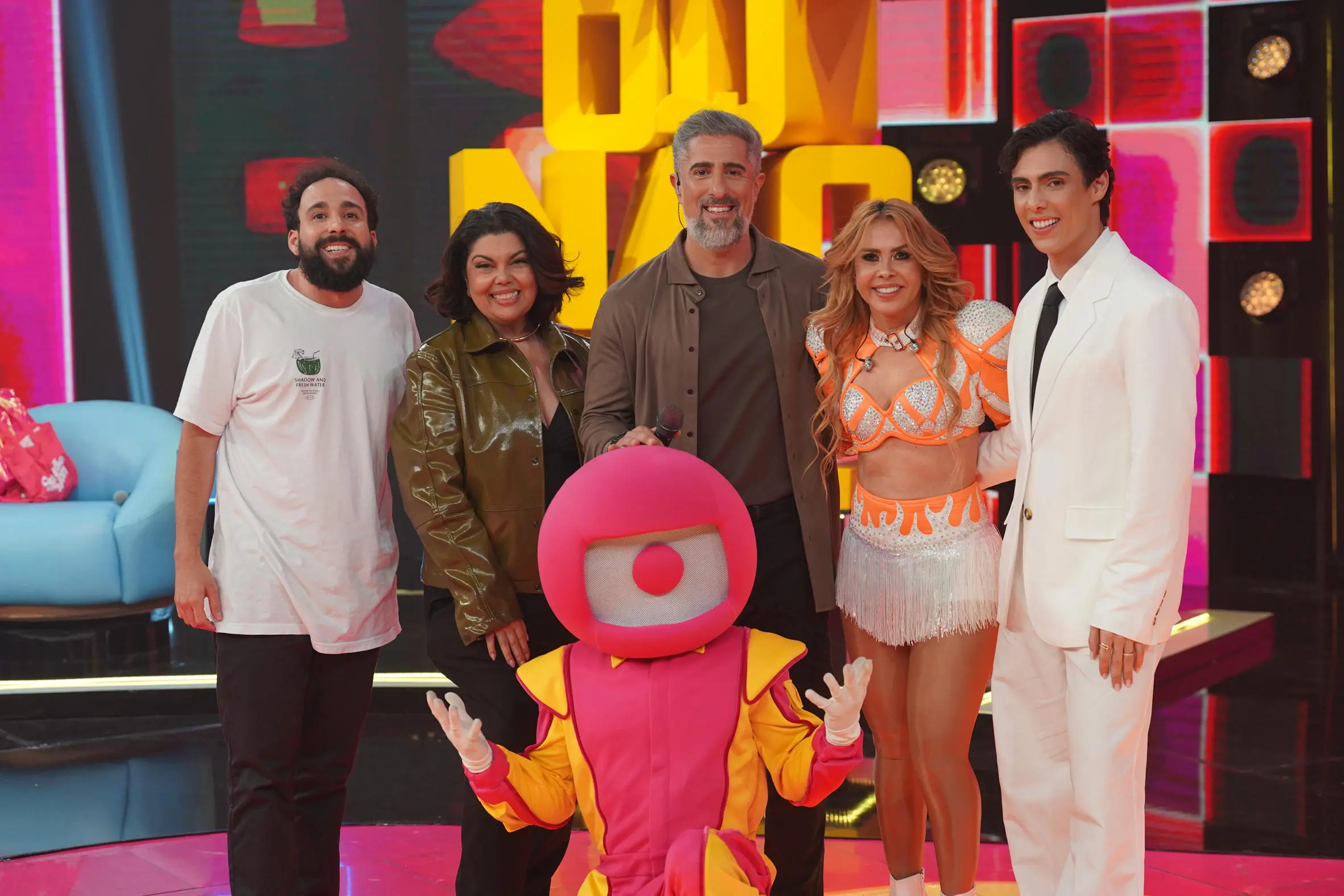 A imagem mostra um grupo de pessoas posando sorridentes em um estúdio colorido, junto com um personagem fantasiado sentado à frente.