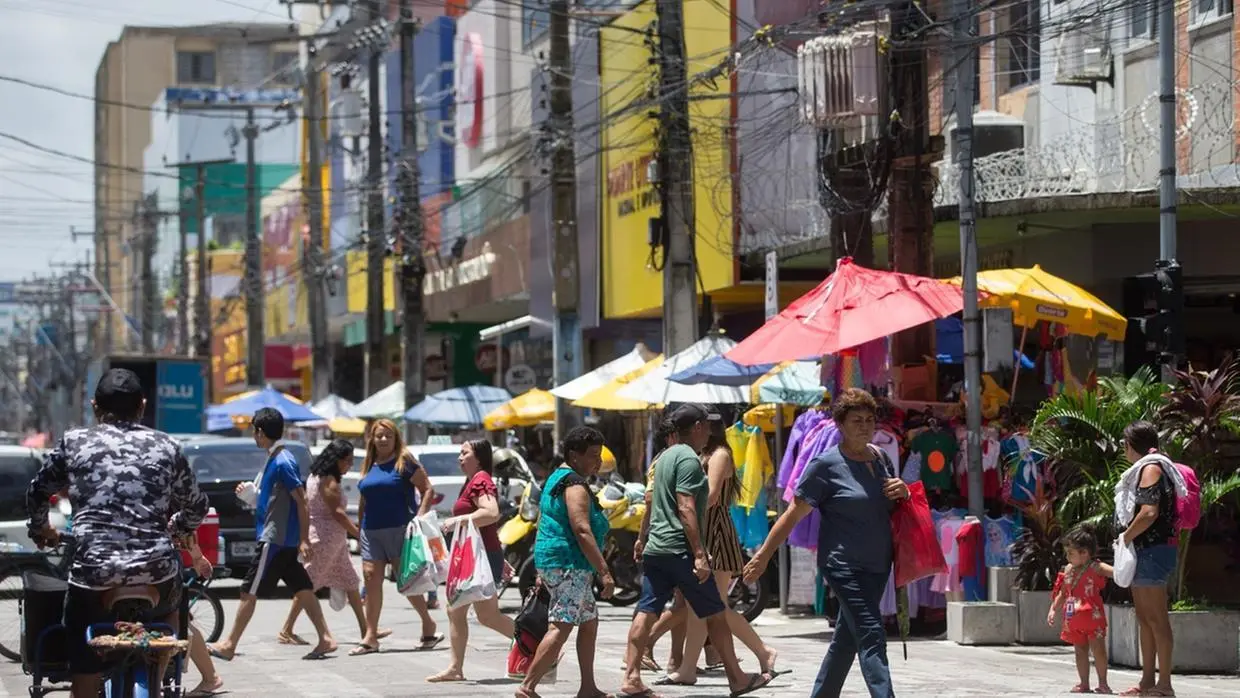 Movimento de pedestres em rua comercial do Ceará, com barracas coloridas, lojas populares e tráfego intenso.