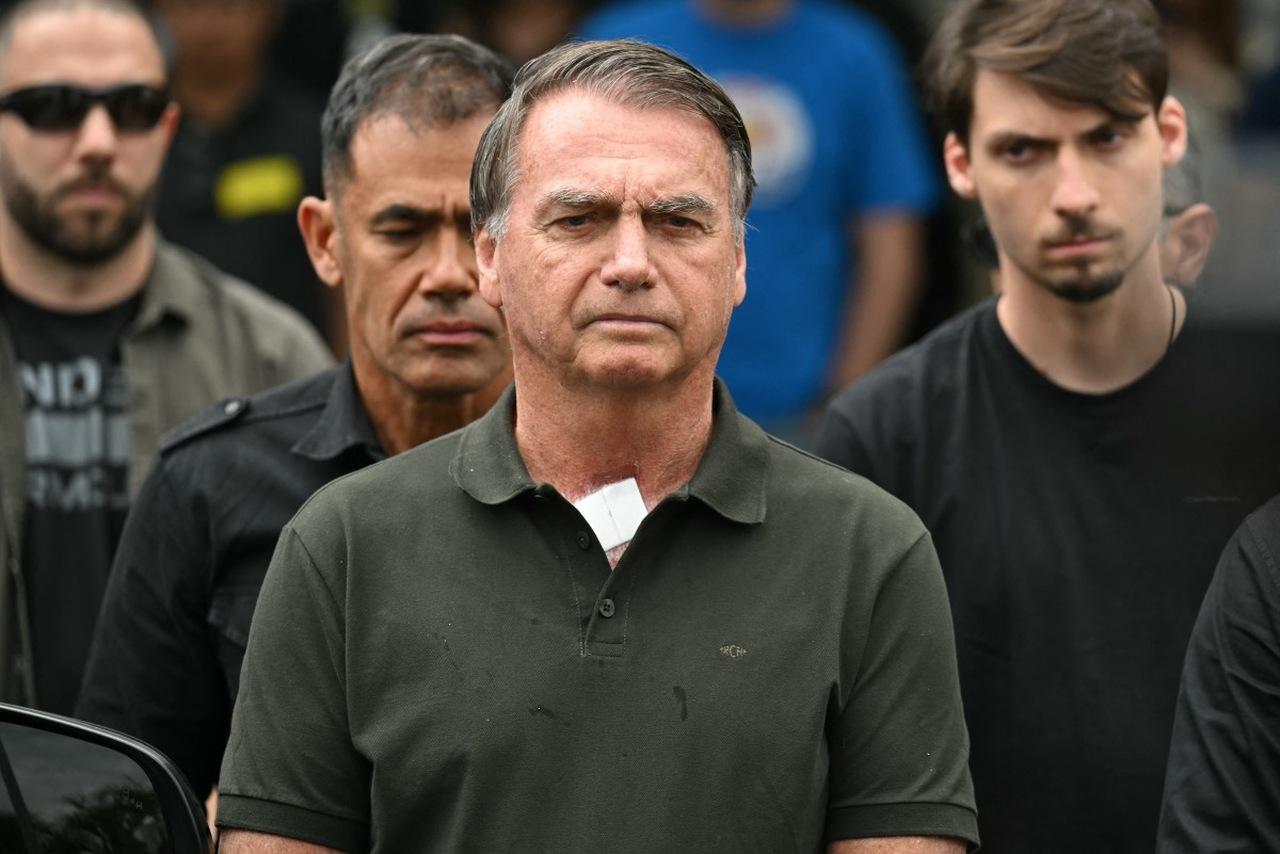 Homem branco de meia idade com cabelos castanhos e grisalhos com blusa verde escura e expressão séria cercado por pessoas ao fundo.