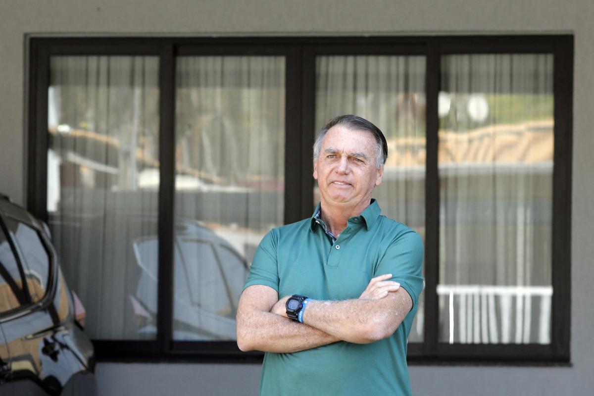 A imagem mostra um homem de meia-idade com os braços cruzados, vestindo uma camisa polo verde. Trata-se de Jair Bolsonaro. Ele está em frente a uma casa, posicionado diante de uma grande janela com cortinas claras. À esquerda, parte de um carro escuro aparece no enquadramento. O homem olha para a câmera com expressão neutra, em um ambiente externo iluminado pela luz do dia.