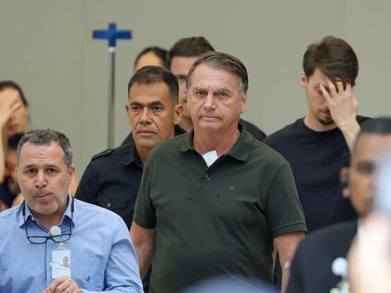 Rosto do ex-presidente Jair Bolsonaro caminhando. Ele aparece de camisa gola polo verde escura em destaque em meio a vários homens que o cercam